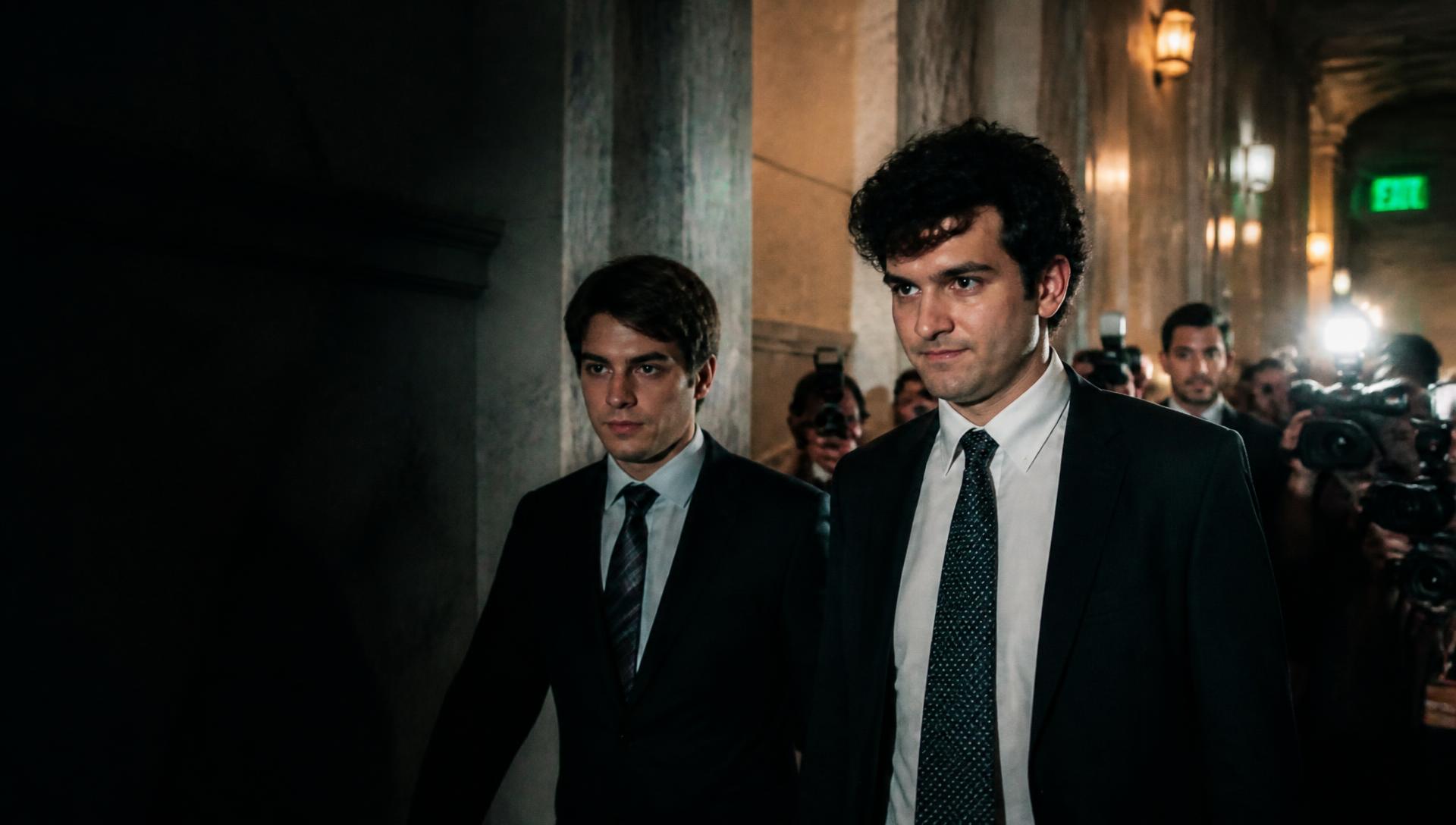 Two founders moving through a crowded government corridor surrounded by press photographers